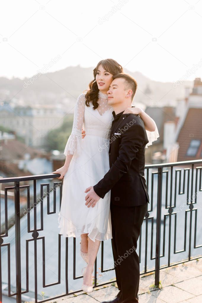 Charming Chinese wedding couple hugging on the terrace with amazing view of old European city architecture