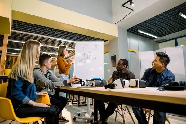 Молодая женщина стоит рядом с белой офисной доской с картой и разговаривает со своими коллегами. Мультирасовая команда людей, проводящих встречи, тестирующих очки виртуальной реальности в офисе, сидящих за столом — стоковое фото