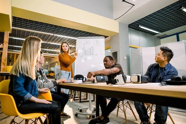 Ung kvinna stående nära White Office Board med diagram och tala med sina kollegor. Multirasistiska team av människor som har möte, testa virtuella verklighet glasögon i kontoret sitter vid bordet — Stockfoto
