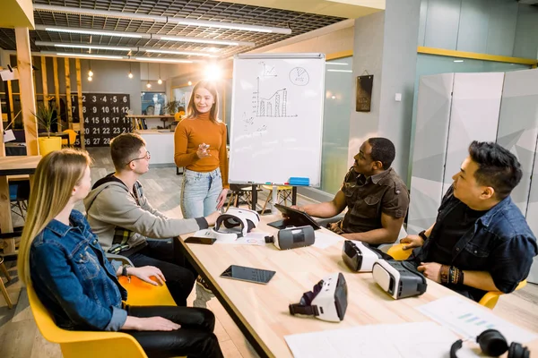 Ung kvinna stående nära White Office Board med diagram och tala med sina kollegor. Multirasistiska team av människor som har möte, testa virtuella verklighet glasögon i kontoret sitter vid bordet — Stockfoto