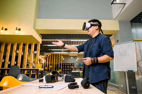 Förvånad asiatisk ung vuxen smart casual man bär virtuell verklighet VR headset gest händer när du står i modern Loft Office — Stockfoto