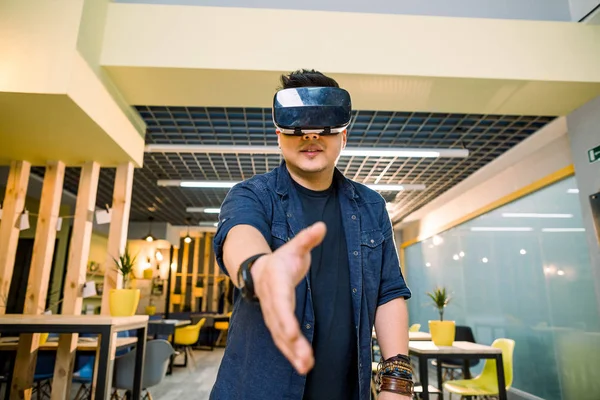 Young Chinese businessman working in office with virtual reality glasses on head. Man using VR headset for handshaking in augmented reality