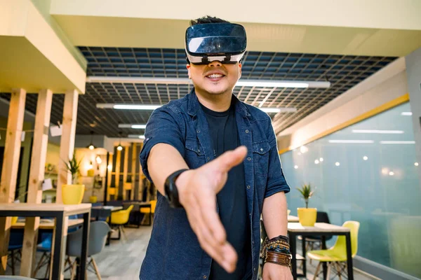 Young Chinese businessman working in office with virtual reality glasses on head. Man using VR headset for handshaking in augmented reality