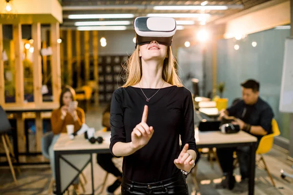 Joyeux jeune femme ajustant son casque de réalité virtuelle et excité de jouer à un jeu vidéo dans l'appareil VR tout en se tenant à son lieu de travail au bureau. Équipe de pigistes sur le fond — Photo