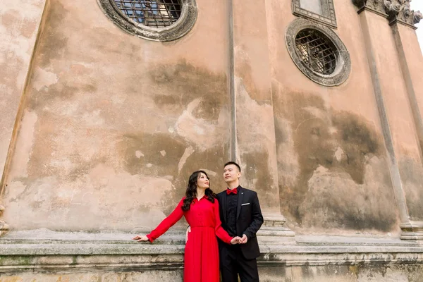 Stylish young Asian couple walking on european old city streets, have fun and hugging, posing near ancient building. Wearing romantic luxury outfit