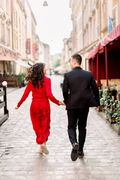 Eski Avrupa şehrinde cadde boyunca eliyle koşan çift. Geri görünüm. Yüzü yok. Takım elbiseli adam ve kırmızı elbiseli kadın. Şehirde aşk — Stok fotoğraf