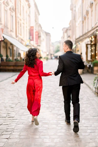 Eski şehir merkezinde cadde boyunca koşarken el ele tutuşan aşık mutlu ve sevimli çiftin geri görünümü. Zarif kırmızı elbiseli kadın ve siyah takım elbiseli erkek — Stok fotoğraf