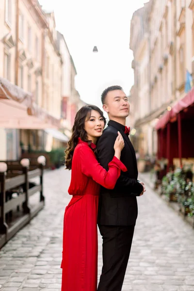 Side view of pretty Chinese woman in red dress keeping hand on shoulder of her man in black suit, and looking away against background of old city buildings.