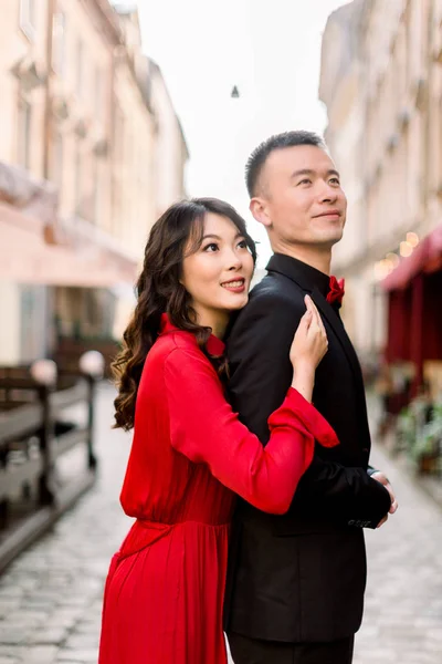 Side view of pretty Chinese woman in red dress keeping hand on shoulder of her man in black suit, and looking away against background of old city buildings.