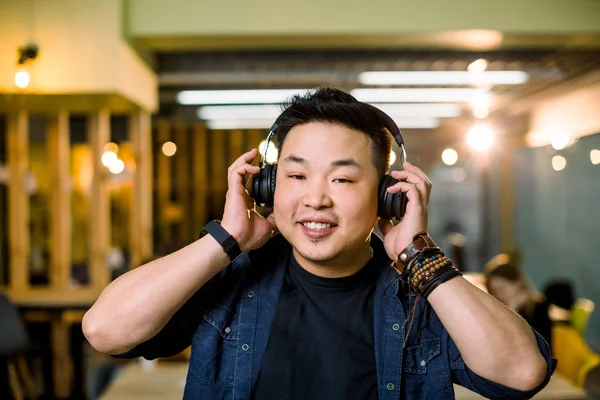 Smiling handsome Chinese man wearing dark blue shirt looking straight ahead while standing in modern office while listening to music over the ear headphones