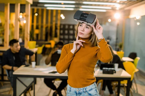 Jolie femme d'affaires en pull orange essayant des lunettes 3d vr au travail. Équipe multiraciale de jeunes travaillant ensemble dans un bureau créatif — Photo