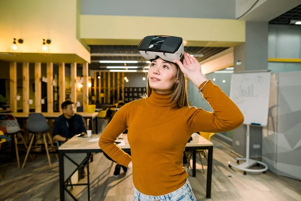Kvinna i orange tröja och jeans bär VR-glasögon bevakade av hennes mångetniska kollegor i modern Office — Stockfoto