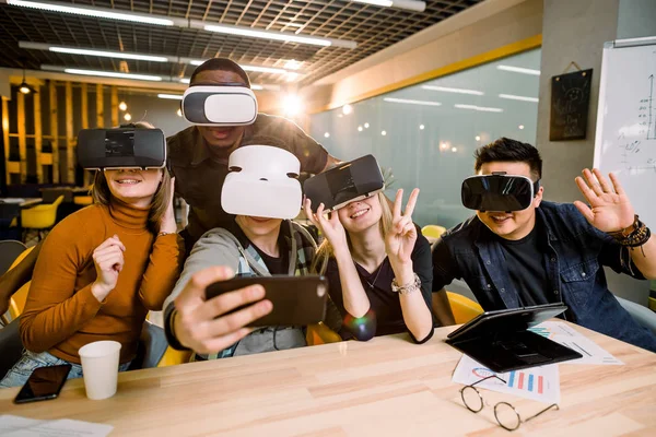 Five young funny people sitting at the table in front of each other, using virtual reality goggles, having fun and gesturing. VR goggles concept, business meeting.
