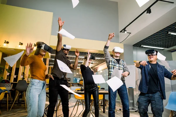 Grupo de jovens empresários multirraciais diversos vestindo óculos VR, jogando papéis para cima, celebrando a sua vitória no escritório loft moderno criativo — Fotografia de Stock