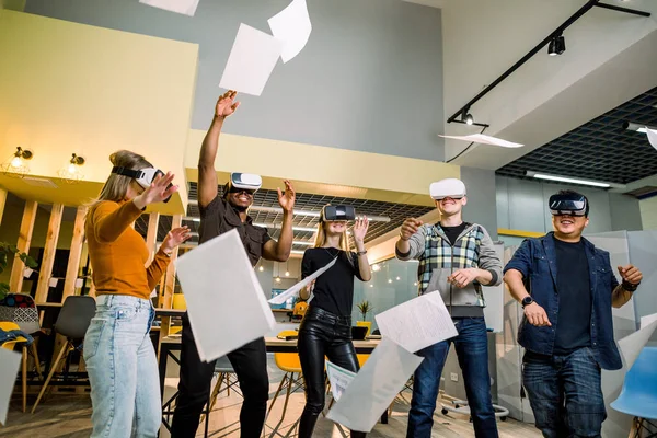 Expresivos cinco jóvenes multiétnicos que llevan gafas de realidad virtual mirando los periódicos mientras están de pie en el moderno estudio de coworking empresarial —  Fotos de Stock