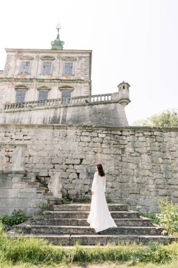 beautiful bride in white wedding dress stands on the stone stairs on the backround of old castle
