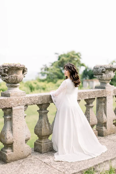 Increíble morena novia asiática china en vestido de novia de lujo, posando en el fondo del castillo viejo, de pie cerca de las barandillas de piedra, foto de la boda, ciudad europea, día de la boda, retrato —  Fotos de Stock