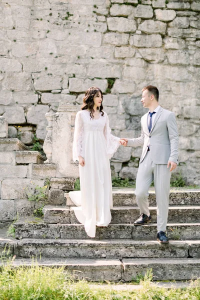 Romantic newlywed Asian couple posing in the ruins of an old castle, holding hands and standing on the stone stairs — Stock Photo, Image