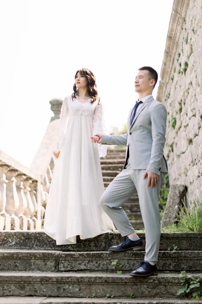 Romantique couple asiatique nouvellement marié posant dans les ruines d'un vieux château, tenant la main et debout sur les escaliers en pierre — Photo