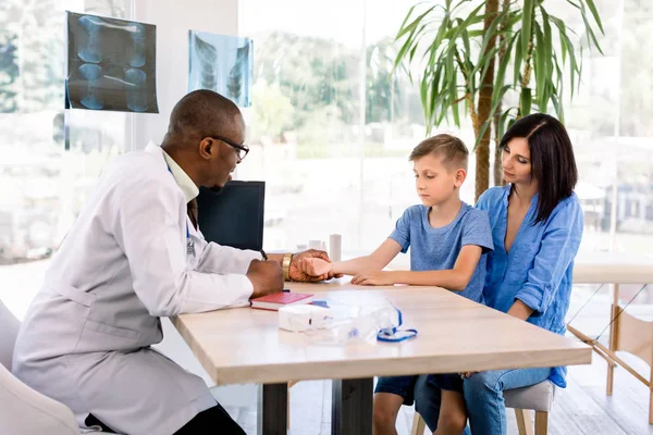 Médico afro-americano pediatra examinando o batimento cardíaco do menino sendo segurado por sua mãe bonita. Clínica moderna, saúde — Fotografia de Stock