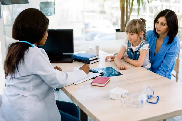 Visão lateral de uma médica afro-americana discutindo sobre o relatório de raio-x com mãe e filha caucasianas no escritório moderno do hospital. Laptop, nebulizador, raio-x na mesa — Fotografia de Stock