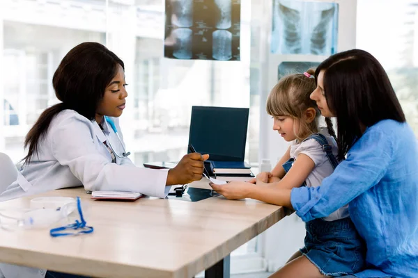 Medicina, cuidados de saúde e conceito de pediatria - mãe muito caucasiana feliz com bebê menina e mulher afro-americana médico na clínica. Eles estão sentados à mesa e conversando — Fotografia de Stock