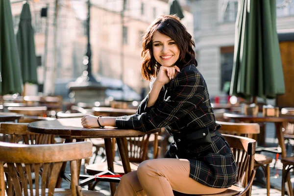 Joven caucásico elegante hermosa mujer en chaqueta negra y pantalones beige, sentado en la silla de madera en la cafetería de la ciudad, al aire libre, estilo de calle, tendencia, elegante — Foto de Stock