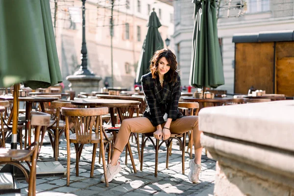 Krásná usměvavá žena ve stylové výbavě, když sedí v kavárně venku, černí a béžové barvy, jarní letní módní trend, elegantní styl, starověké městské budovy na pozadí — Stock fotografie