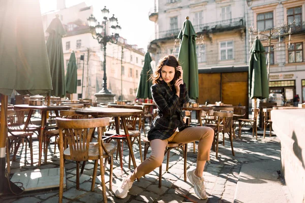 Mulher sorridente bonita na roupa elegante posando enquanto sentado no café ao ar livre, cores pretas e bege, tendência de moda primavera verão, estilo elegante, edifícios da cidade antiga no fundo — Fotografia de Stock