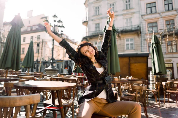 Mooie vrouw zittend in een café op een straat in de stad, mode stijl, handen omhoog, wakker in de ochtend, genieten van zonnige dag — Stockfoto