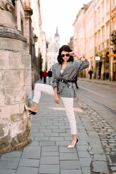 Giovane donna bruna in eleganti abiti da lavoro e occhiali da sole alla moda, posa sexy vicino al vecchio muro di pietra dell'edificio, colpo all'aperto — Foto Stock