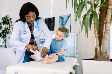 Bandaj koyuyor. Koyu tenli kadın çocuk doktoru beyaz ceket giyen küçük çocuğun bacağında bandaj koyarak