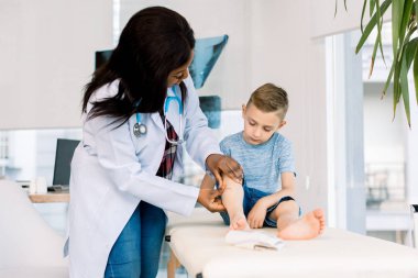 Beyaz önlüklü Afrikalı kadın doktor dikkatle ilk yardım sağlar. Erkek bacağını inceliyor. Çocuk sabırla kanepede otururken, süreci gözlemler.