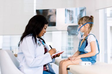 Modern hastane veya klinikte tıbbi kanepede oturan küçük sevimli kız nebülizör ile teneffüs yapan Dostu Afrikalı kadın doktor