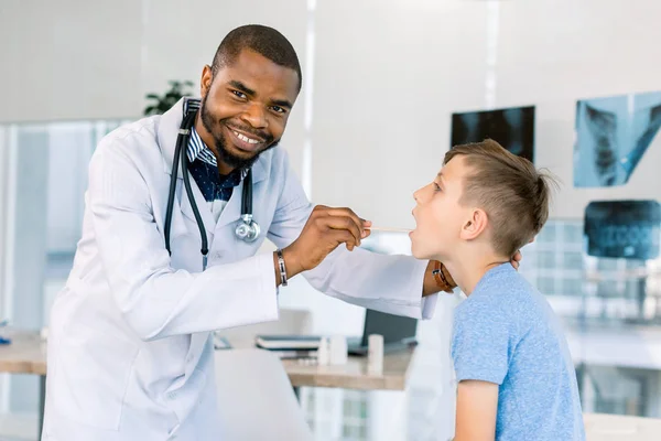 Lycklig afrikansk amerikansk manlig läkare tittar patient pojke med halsont i kliniken. Läkaren undersöker små pojkar halsen på sjukhuset — Stockfoto