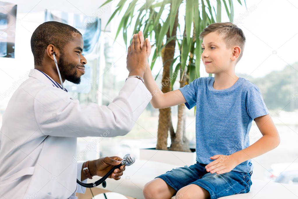 Happy smiling African American male doctor giving five to a little boy in modern medical clinic. Medical examination is fine