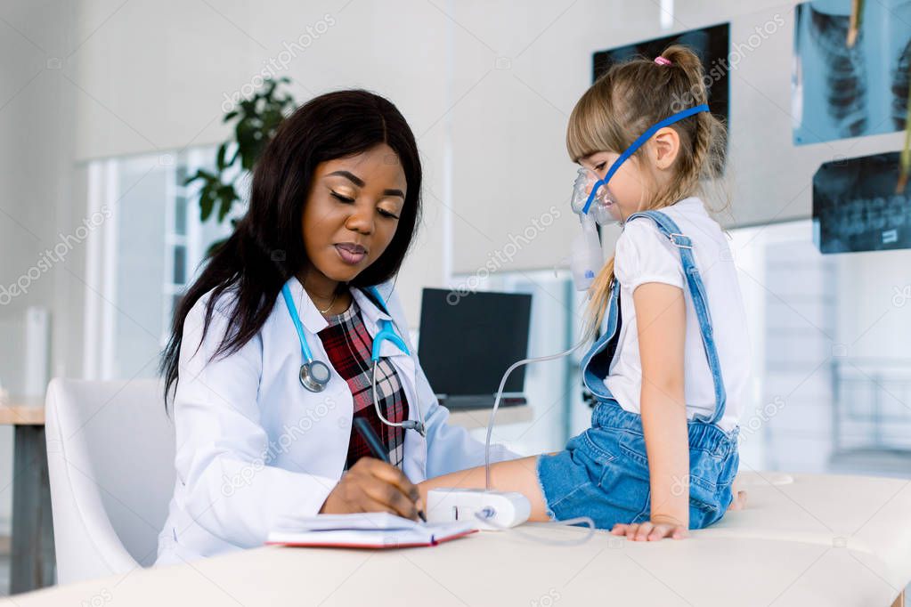 Young African female Doctor helping to little girl with nebulizer mask. Medical doctor applying medicine inhalation treatment on a little girl with asthma inhalation therapy by the mask of inhaler.