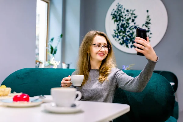 Picture of cute young lady sitting in cafe and making selfie with cup of coffee. Look at phone.