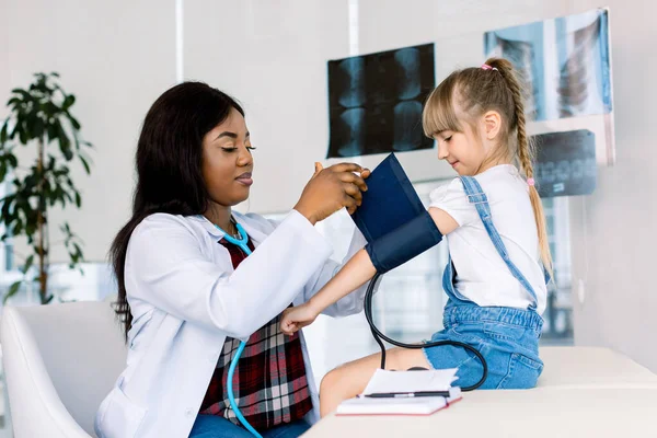Menina visitando jovem médica africana no hospital. Medição da pressão arterial e verificação do pulso — Fotografia de Stock