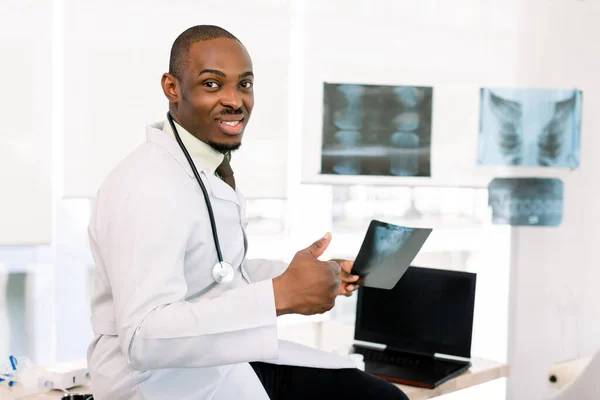 Good news. Cheerful African doctor showing his thumb up and smiling while sitting at his table with computer and holding radiography, against background of modern light medical clinic