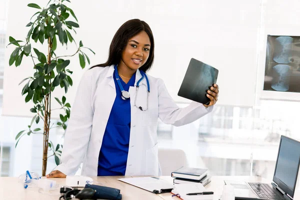 Young confident smiling African American female doctor standing at the table and holding radiography, medical background — 스톡 사진