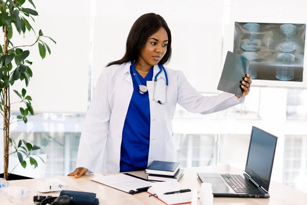 Beautiful female african medical worker holding x-ray, while workong at the table with laptop, books, in modern medical office — 스톡 사진