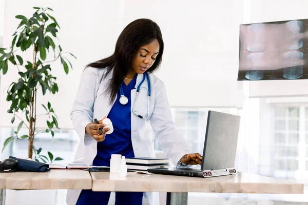 Femme médecin noire se tient à un bureau à la recherche d'un ordinateur portable, infirmière afro-américaine travail sur ordinateur portable dans l'armoire, hôpital moderne — Photo