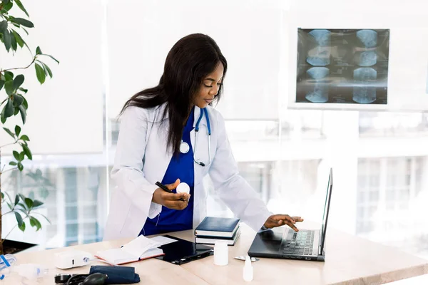 Vue latérale du jeune médecin afro-américain tenant une bouteille de médicament et utilisant un ordinateur portable tout en travaillant au bureau à l'hôpital — Photo
