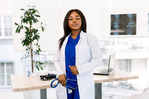 Female pretty African doctor in white coat looking to camera in an modern medical office