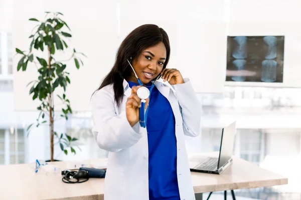 Cute afro-american doctor at her office, holding stethoscope, standing in light office in clinic — 스톡 사진