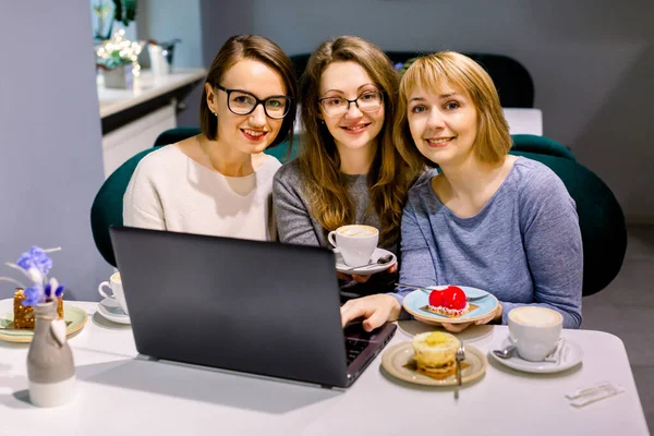Женщины-друзья смотрят в кафе на камеру, едят вкусные десерты и пьют чай, используют ноутбук для работы. Женщины-блогеры-фрилансеры, работающие в кафе — стоковое фото