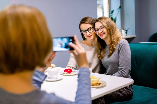 Women friends in cafe indoors. Two pretty women friends hugging and posing for the photo together, while her third friend woman is taking a picture on the smartphone