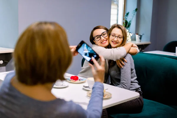 Women friends in cafe indoors. Two pretty women friends hugging and posing for the photo together, while her third friend woman is taking a picture on the smartphone