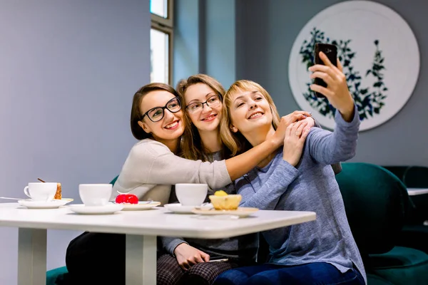 Tres hermosas mujeres jóvenes disfrutando del café y la torta juntas en un café sentado a la mesa riendo y haciendo fotos de selfies con sonrisas felices —  Fotos de Stock